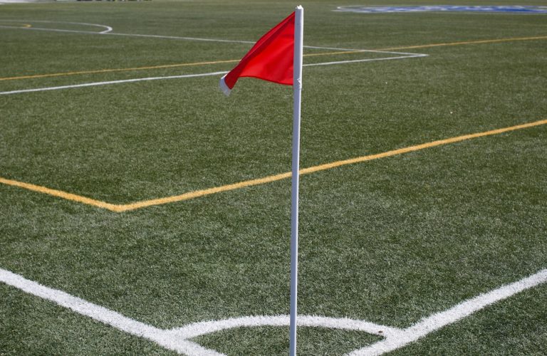 A red and white corner kick flag on a green soccer pitch during daytime.