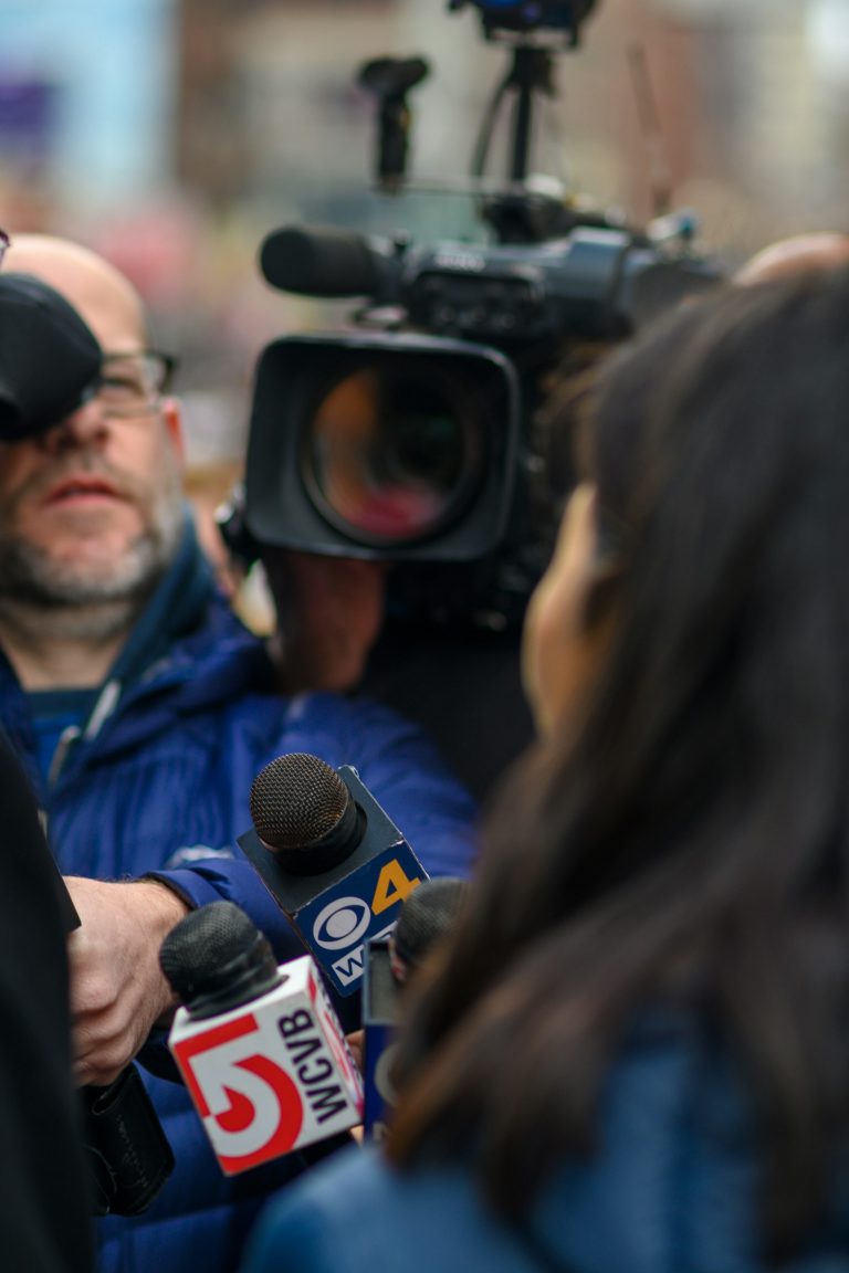 A man surrounded by reporters and microphones at a press conference, used to illustrate the modern beat-writing environment where analytics has reshaped daily coverage workflows.
