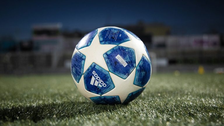 Close-up of an official blue and white Adidas tournament soccer ball resting on stadium grass at dusk, suggesting the evening of a World Cup match