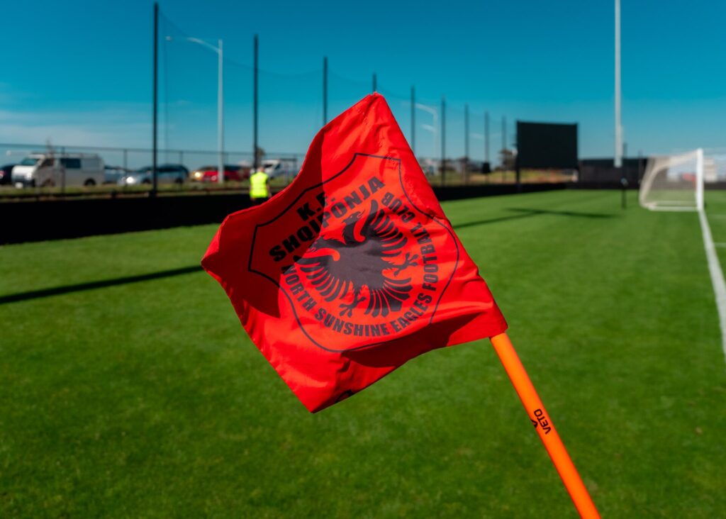 A corner flag in the foreground with players spread across the penalty area behind it